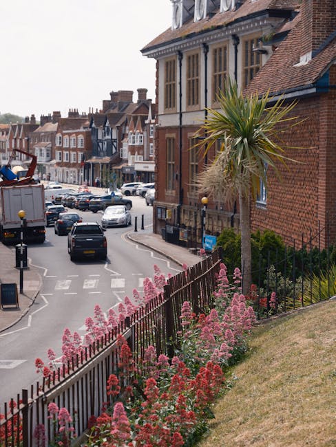 In the foreground, a small garden area features a variety of pink and red flowering plants along a black metal fence, with a tall, spiky palm tree situated nearby. The garden is located on a gentle sl